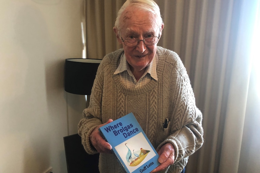 An elderly man with glasses in a white jumper holds a copy of his book with a picture of a brolga on the front in a hotel room