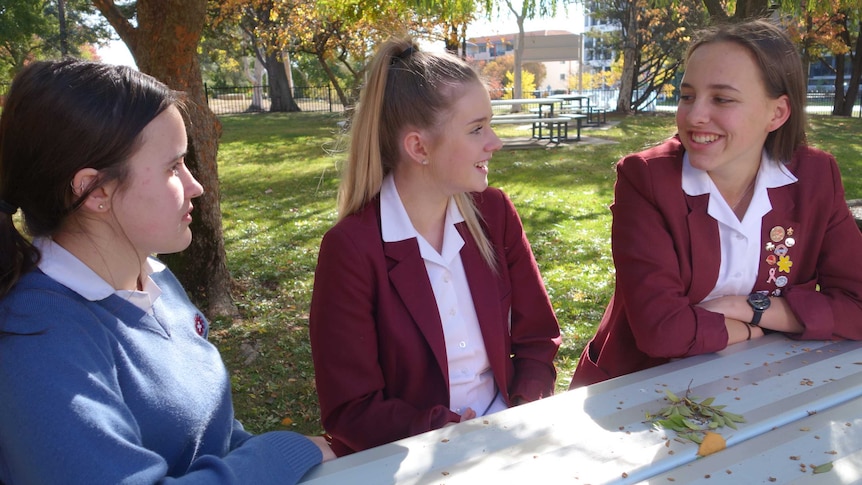Three high school students sitting in the school garden at a table smiling at each other