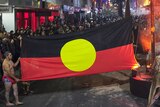 At night in a Hobart street, a young Aboriginal man holds up a Aboriginal flag, a large crowd of people gather behind the flag