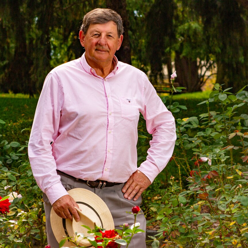 A man with short brown hair, wearing a pink shirt, standing in front of rose bushes and trees.