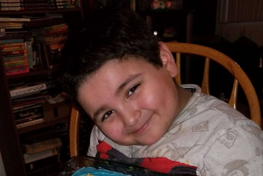 A boy with black hair smiles as he bends over a birthday cake of an elephant sitting on grass.