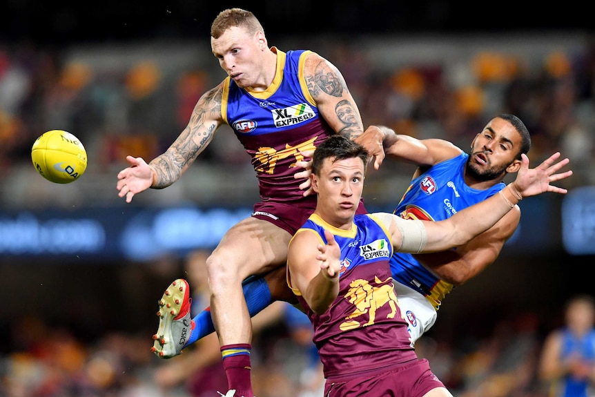 Two Brisbane Lions AFL players contest for the ball in the air alongside a Gold Coast Suns opponent.