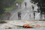 People with umbrellas watch from far away as debris swirls in floodwaters on a road