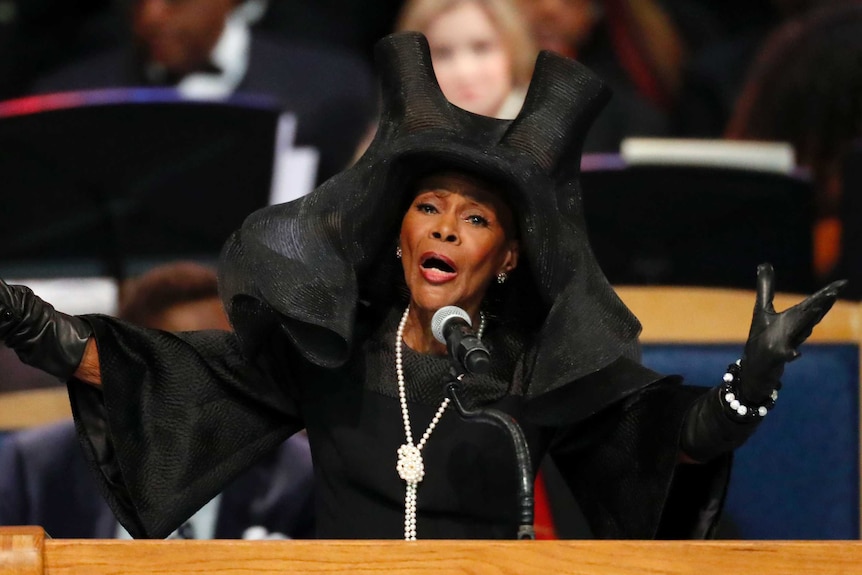 Cicely Tyson wears a big black hat and what appears to be a black dress with a pearl necklace on top. She wears leather gloves.
