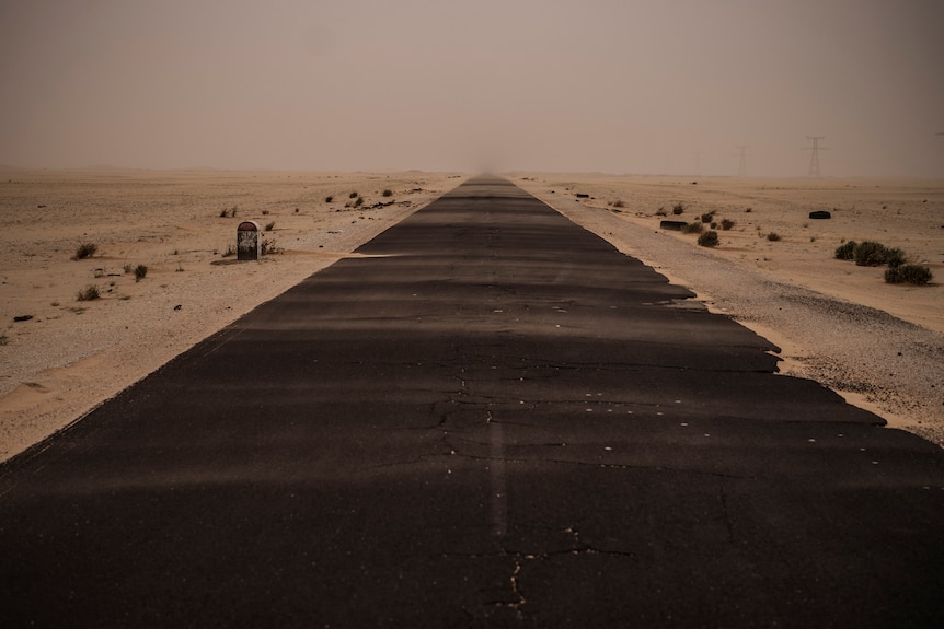 An image from the middle of a strip of tarmac leading through a dusty, dry and sandy environment.