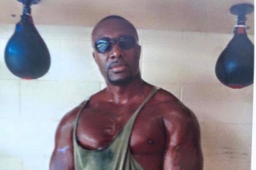 A dark skinned buff man stands facing the camera with glasses on