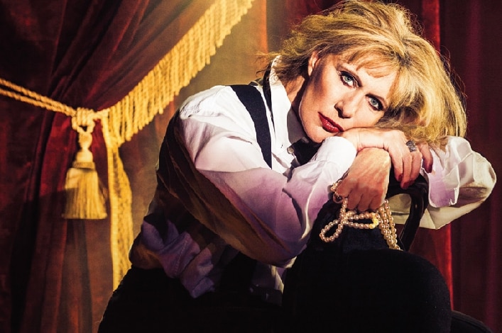 woman with blonde hair white shirt and black waistcoat resting head on forearms on back of a chair in front of red curtain