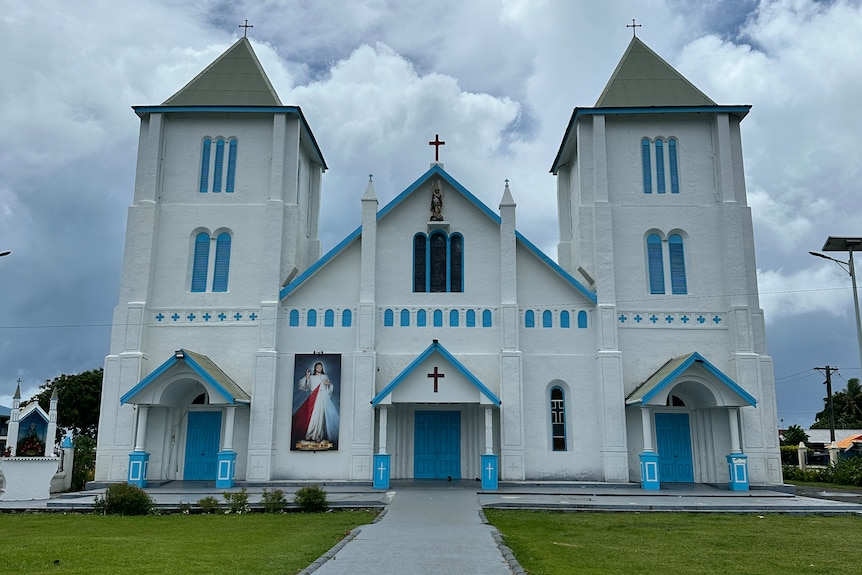 The white facade of St Michael Archangel Catholic Church, Leauvaa, with a picture of Jesus and light blue trims.
