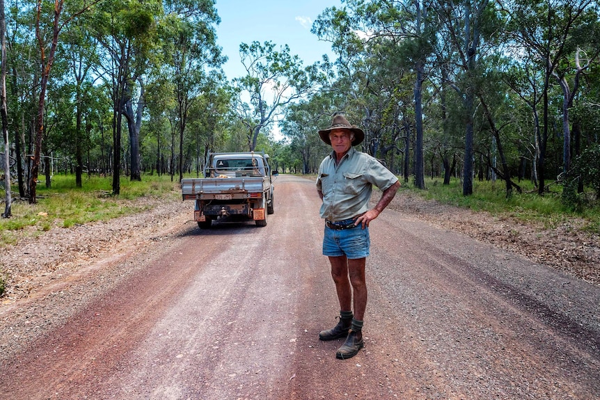 A man in an Akubra-style hat, shirt and shorts, stands with his hands on his hips near a ute on a dirt road surrounded by trees.