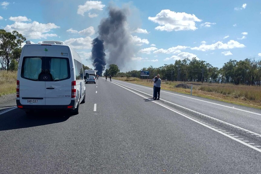 People stand next to a van on a highway. A large plume of smoke billows high in the distance.