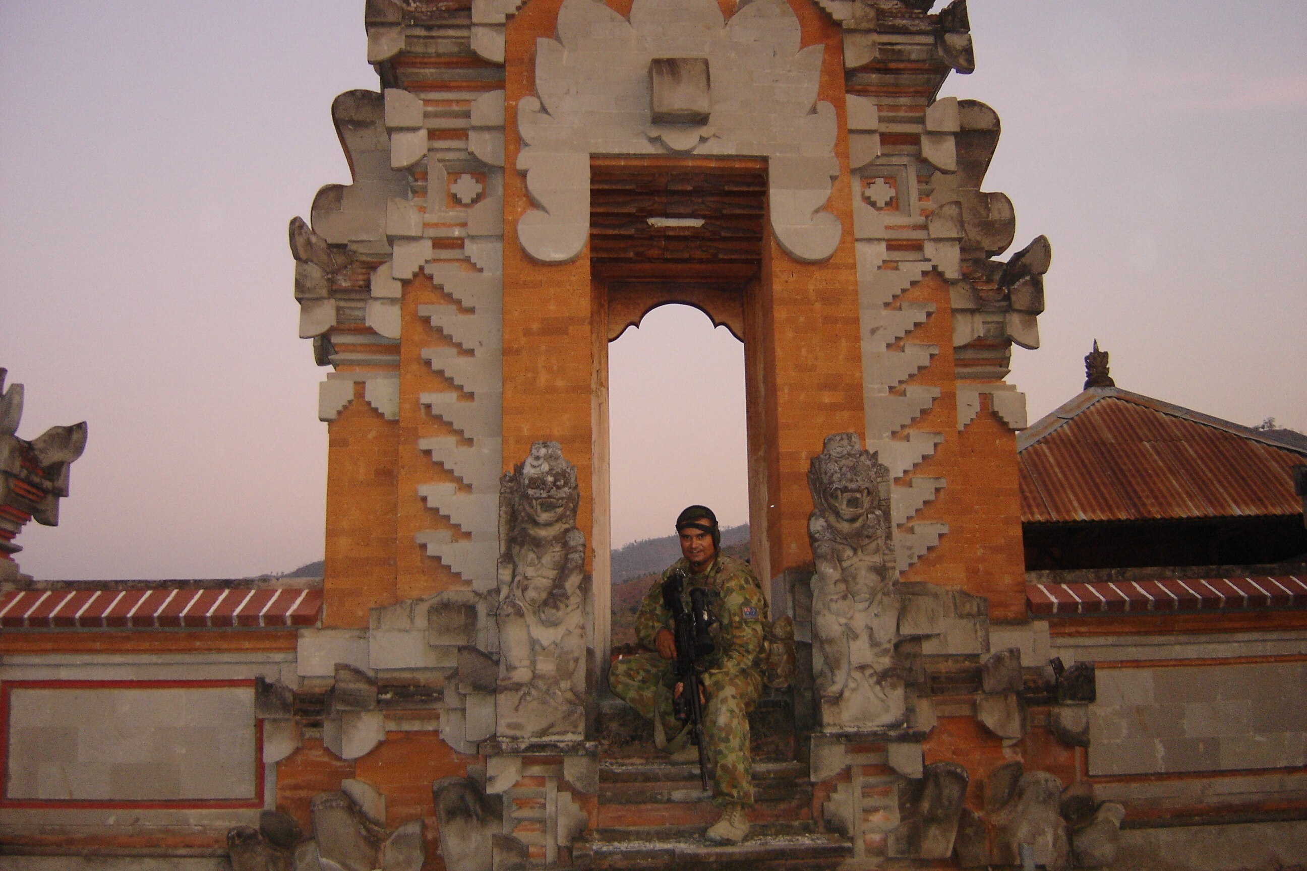 A soldier poses for a photo on the steps of what appears to be a temple with a nice sky in the background.