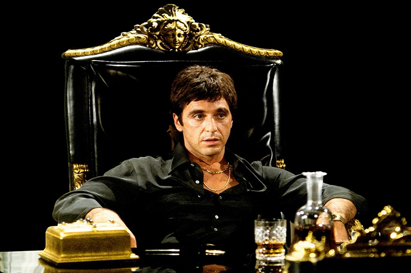 A man with short brown hair wears gold chains and black button up shirt and sits in ornate black and gold chair at desk.