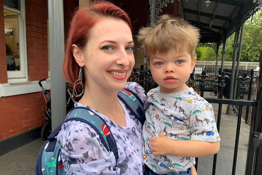 Lina Gyle, wearing a child's backpack, holds her son outside a redbrick building.