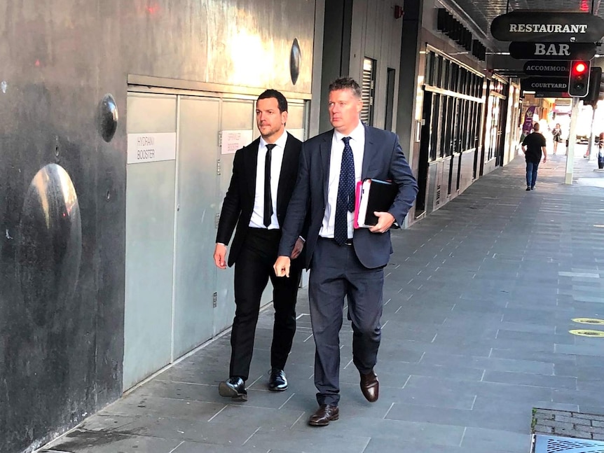 Jarrod Mullen wearing a black suit and tie walking down Newcastle's Hunter Street with his lawyer.