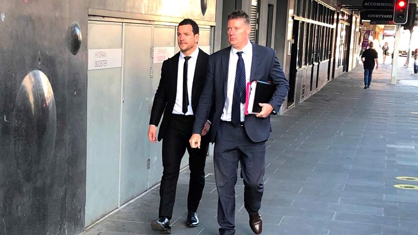 Jarrod Mullen wearing a black suit and tie walking down Newcastle's Hunter Street with his lawyer.