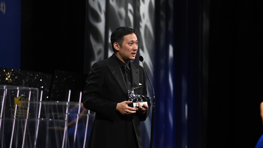 A man in a suit holds a silver trophy and speaks into a microphone on a stage.