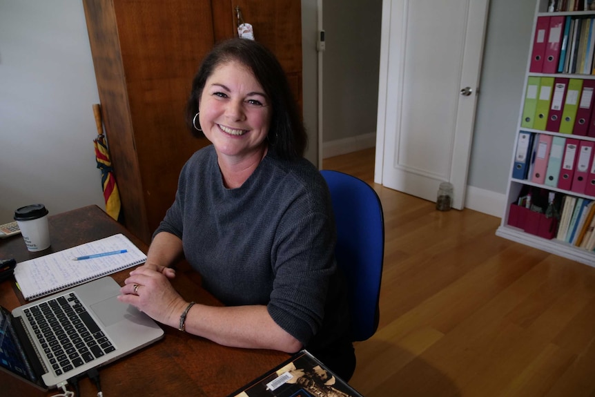 A woman with dark hair sits at a desk on a computer smiling, in a home office.
