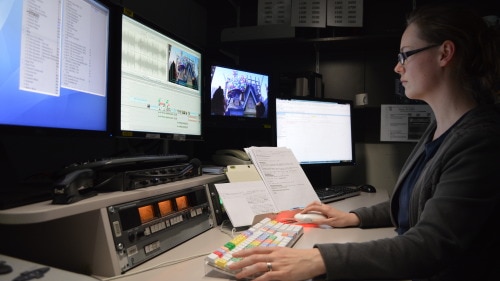 A lady with glasses sits at three computer screens editing.