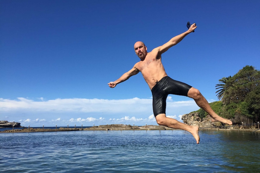 A man in swim shorts pulls a pose and a funny face mid air as he jumps from the edge into an ocean pool.