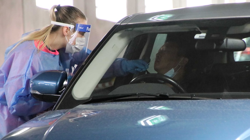 A healthcare worker wearing a blue gown and face shield sticks a swab up the nose of a man driving a Range Rover.