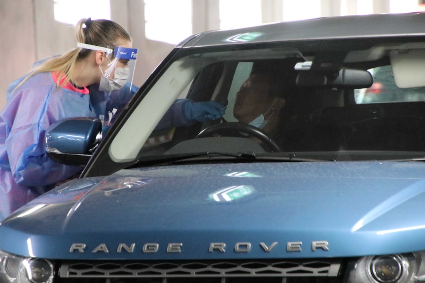 A healthcare worker wearing a blue gown and face shield sticks a swab up the nose of a man driving a Range Rover.