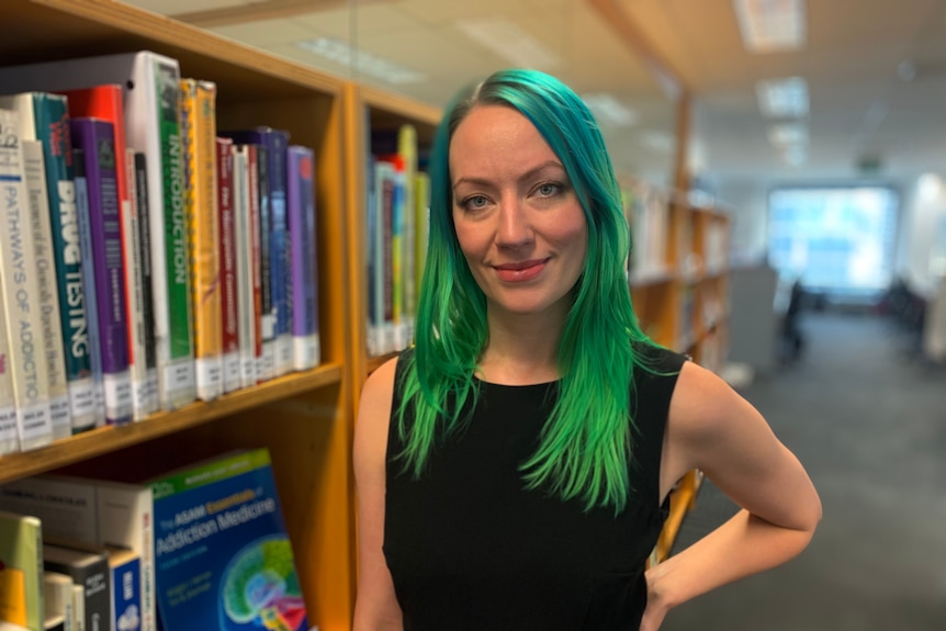 A woman with vivid teal hair stands next to a book case.