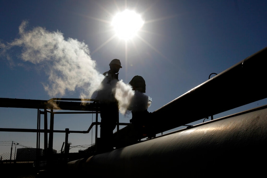 An oil employee is backlit against a harsh low sun, as they stand atop oil refinery pipes on a clear day.