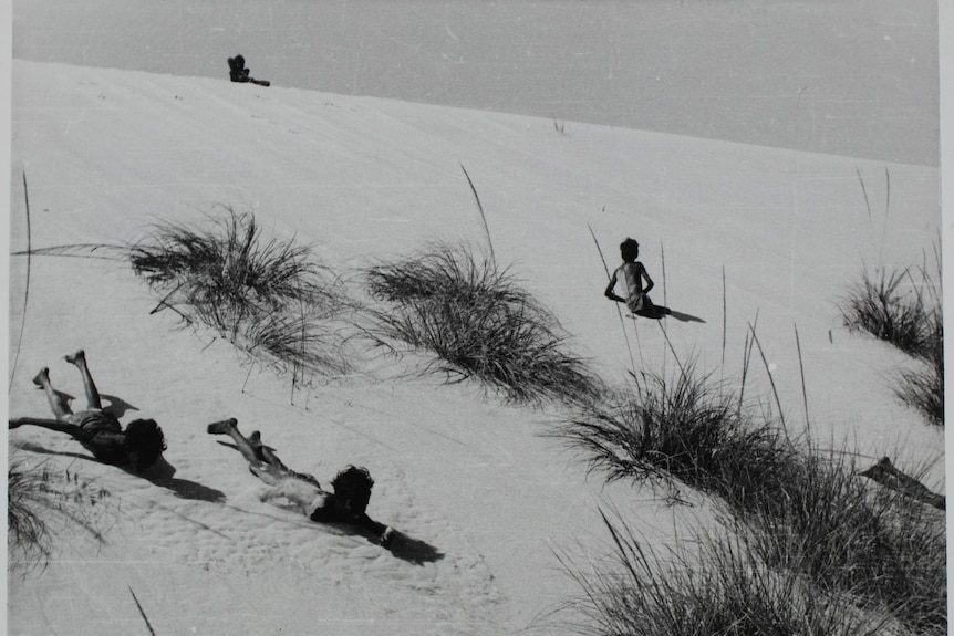 Three boys sliding down a sand dune on their bellies.