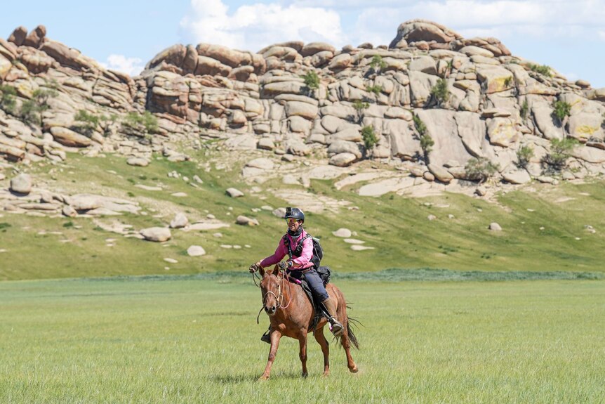 A woman races a brown horse across the Mongolian savannah, with rocks in the background.