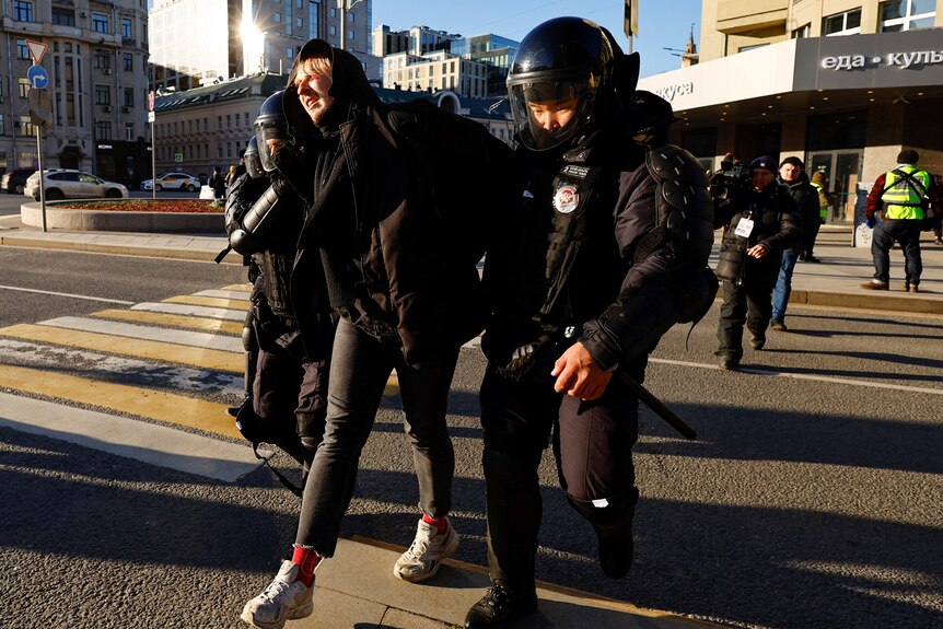 A man with blonde hair is held by Russian police as they walk across a street.