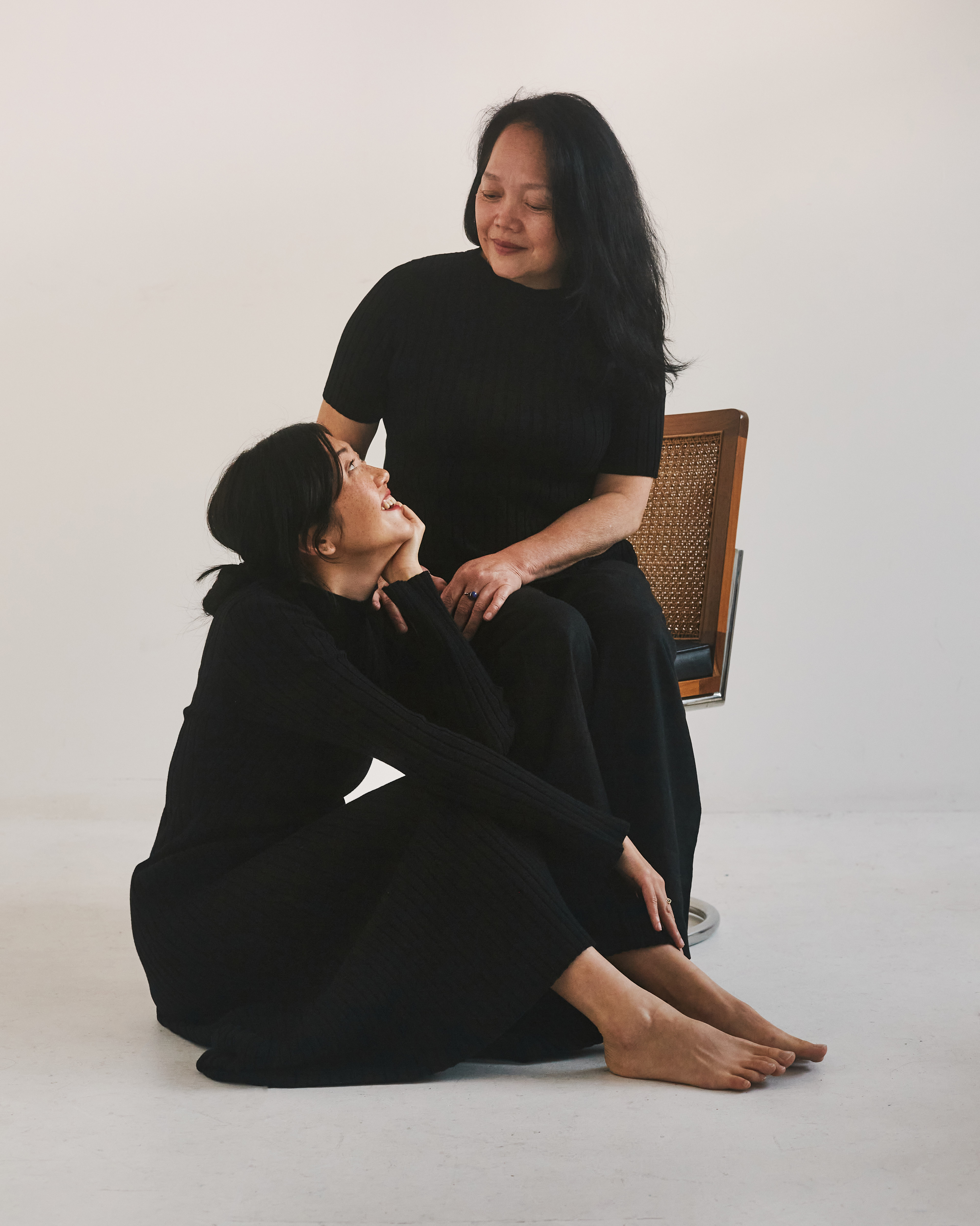 Rainbow, a 30-something Chinese Australian, sits at her mother Irene's feet and smiles up at her. Both are wearing all black.