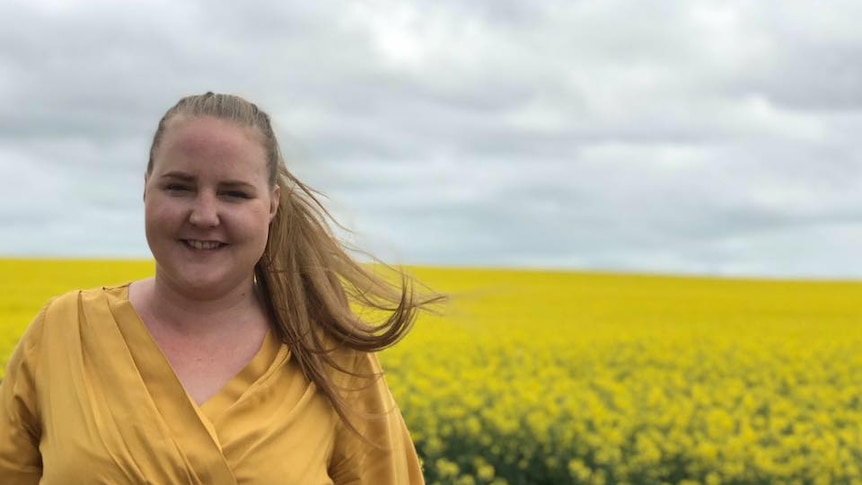 A smiling woman wearing a yellow top stands in front of a field of yellow flowers