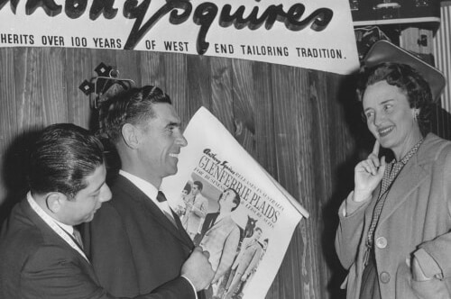 A black and white photo showing two men in suits looking at a photo, with a woman looking on.