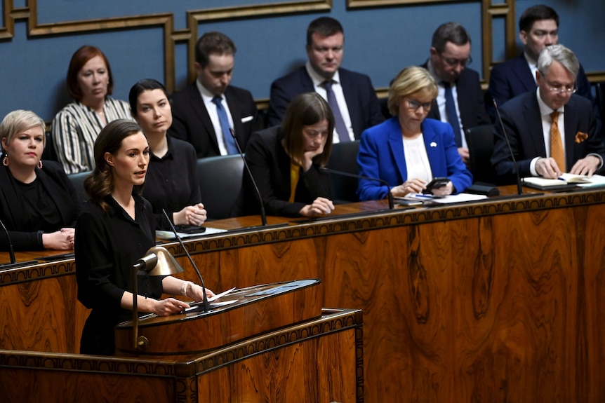 Une jeune femme aux cheveux bruns s'adresse au Parlement finlandais depuis un podium en bois d'apparence riche.