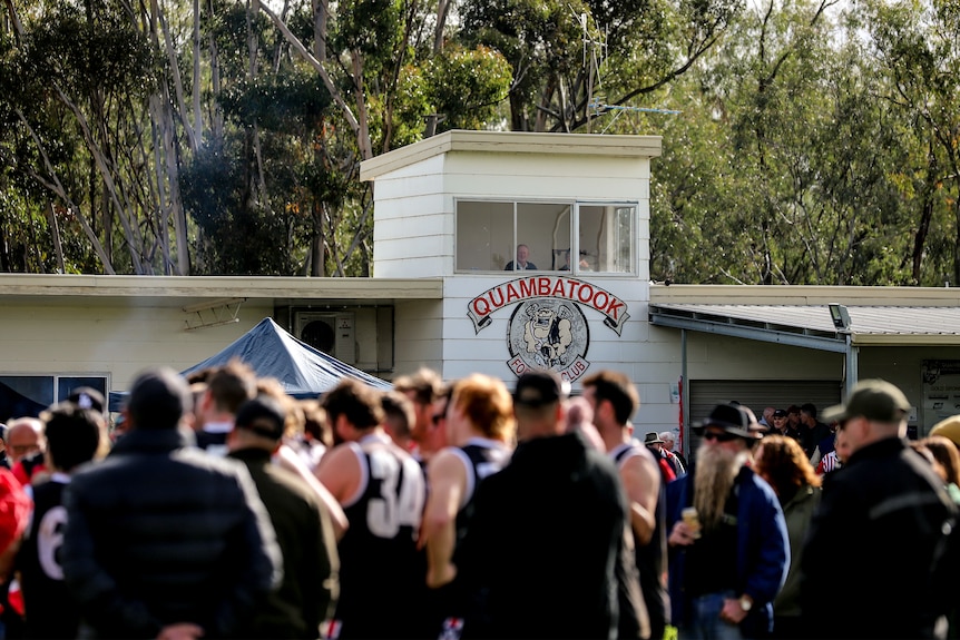 A man sits in a raised timber grandstand watching over players and spectators on a country football oval