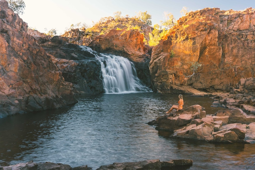 A woman in swimsuit sitting on a rock watching the waterfall at Edith Falls at sunrise