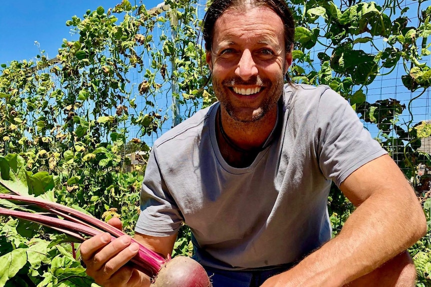 Smiling man crouching in a garden, holding a large, freshly picked beetroot