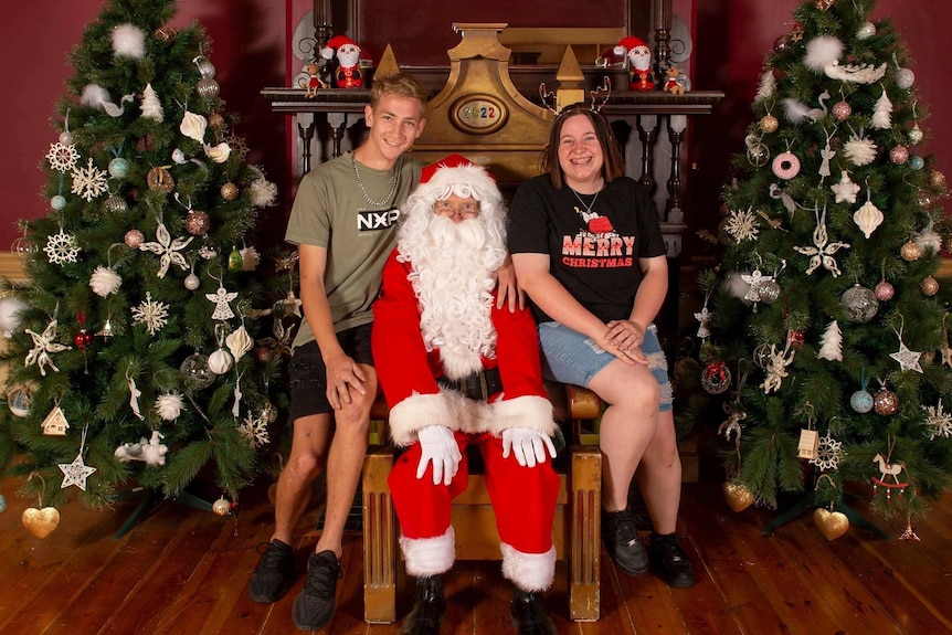 A man and a woman sitting next to Santa in the middle of two Christmas trees