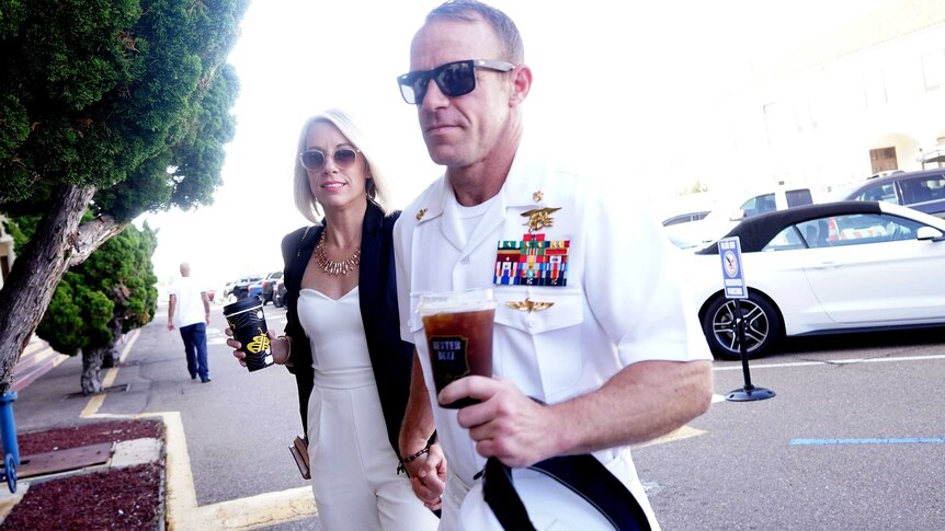 man in white navy uniform, lots of pinned medals, carrying take away coffee and holding hands with blonde-haired man