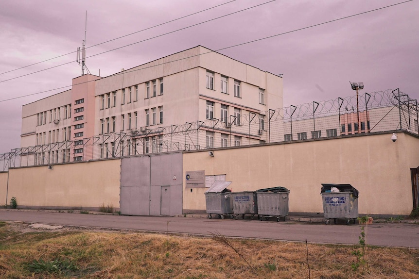 The exterior of the Busmantsi Detention Centre is seen, with dark grey skies overhead. The building is yellow and grey.
