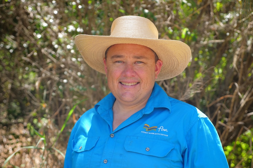 A man in a hat a bright blue shirt smiling in front of some trees and shrubs.