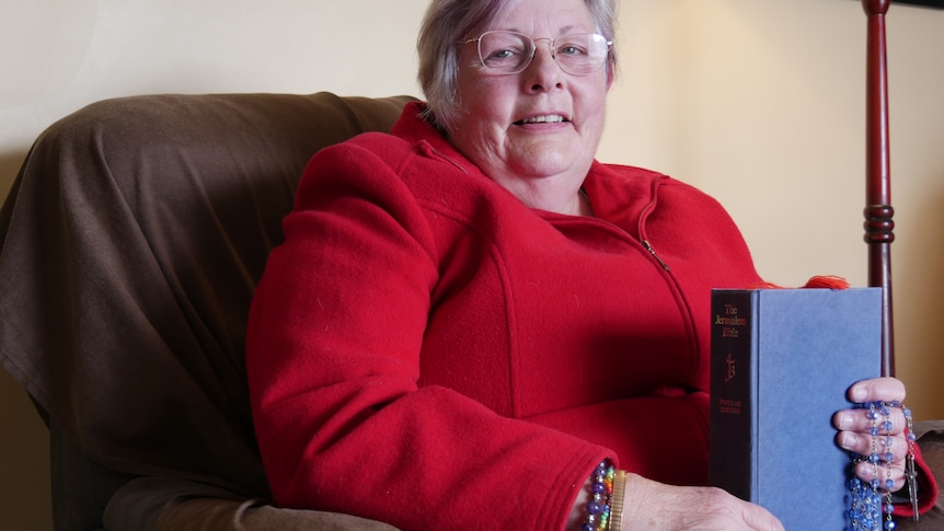 A woman in a red jacket sits brightly in a chair, clutching a blue bible and colourful dark blue rosary beads