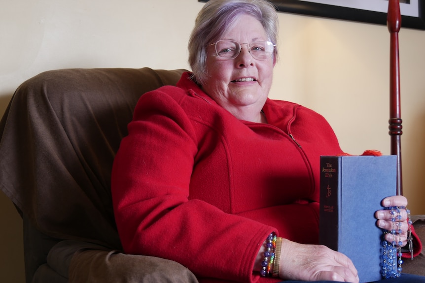 A woman in a red jacket sits brightly in a chair, clutching a blue bible and colourful dark blue rosary beads