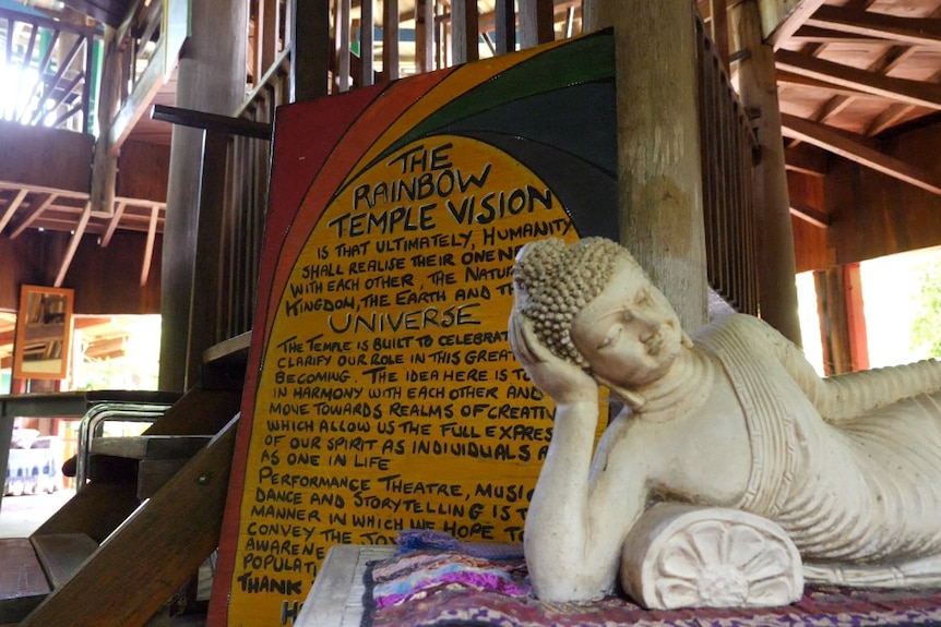 The inside of a wooden building with stairs, a yellow sign and a buddha statue in the foreground