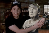 A film still from Wayne's World, of a man in a cap, hugs a Roman-style bust of Donald Trump