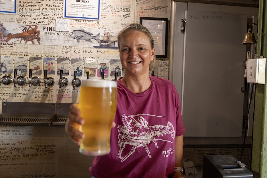 Woman holds beer in pint glass towards camera as she smiles. 