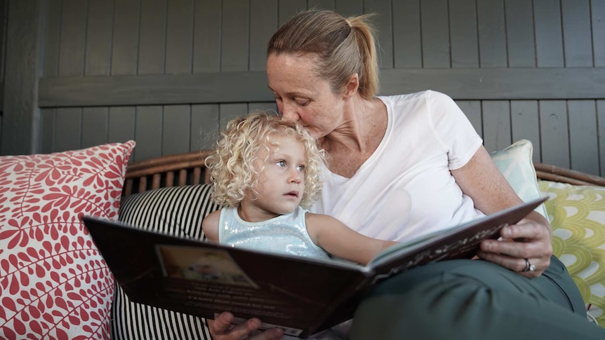 A woman and a small child sit on an outdoor lounge with a large book.