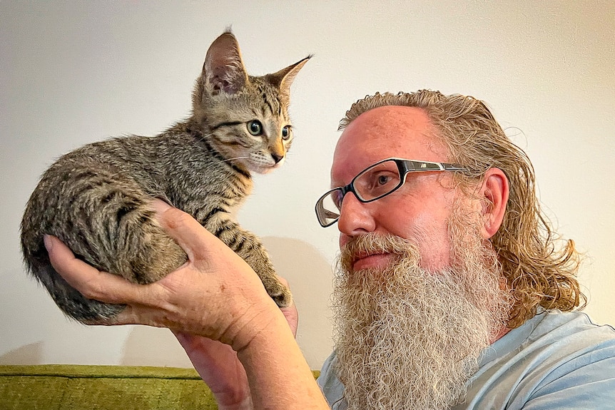 Bearded, long-haired man holding up kitten while sitting on couch.