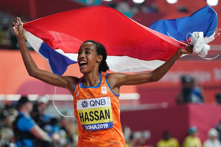 Sifan Hassan carries a blue, red and white flag in both hands above her head with her mouth open wearing an orange singlet