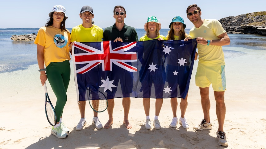 A group of Australian tennis players stand on a beach, some holding racquets, with an Australian flag in front of them.  